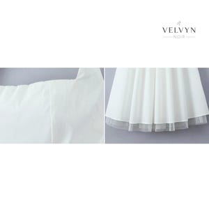 May include: A white dress with a layered skirt and a sheer hem. The dress has a simple, elegant design, perfect for special occasions. The brand name "VELVYN NOIR" is displayed in the upper right corner.