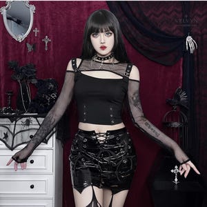 May include: A black cropped top featuring long fishnet sleeves, a mesh chest panel, and silver-toned buckle straps at the shoulders. Paired with a black patent leather mini skirt that includes a front lace-up closure and silver metal eyelets.
