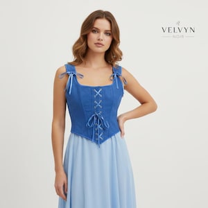 K&ouml;nnte beinhalten: Ein blaues Jeans-Korsett-Top mit Schn&uuml;rung vorne und Bindetr&auml;gern. Das Top wird mit einem hellblauen Rock kombiniert. Der Markenname "VELVYN NOIR" wird oben rechts angezeigt.