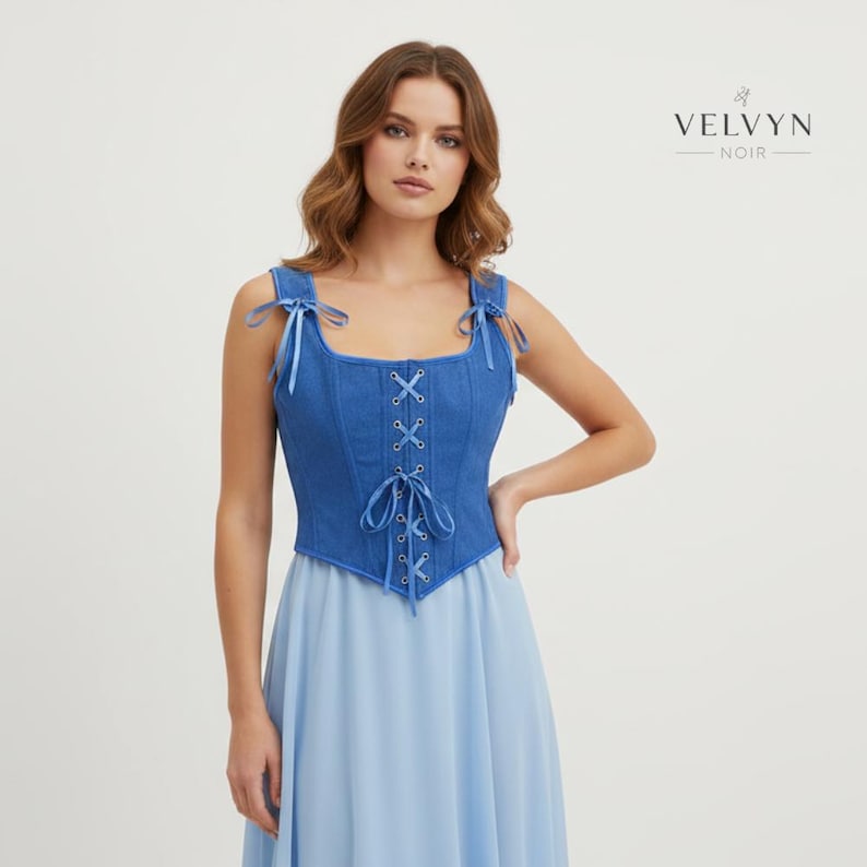 K&ouml;nnte beinhalten: Ein blaues Jeans-Korsett-Top mit Schn&uuml;rung vorne und Bindetr&auml;gern. Das Top wird mit einem hellblauen Rock kombiniert. Der Markenname "VELVYN NOIR" wird oben rechts angezeigt.