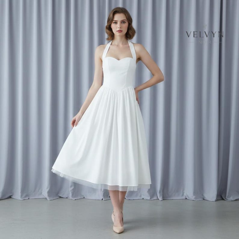 May include: A white halter-neck dress with a sweetheart neckline and a flowing skirt. The dress has a fitted bodice and a tea-length hemline. The model is wearing nude-colored heels. The brand name "VELVYN" is visible in the upper right corner.