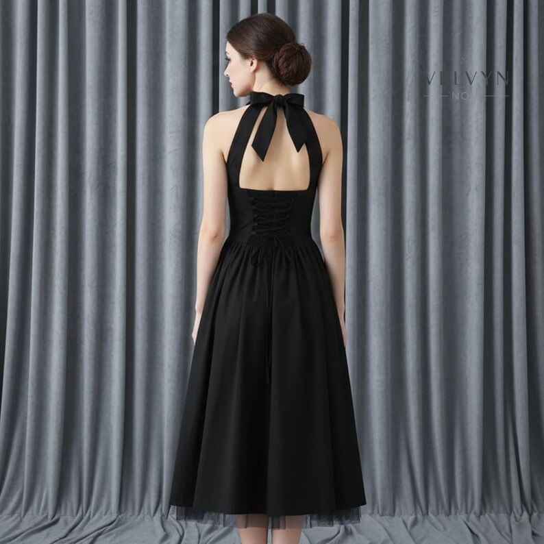 May include: A black halter dress with a bow at the neck and a corset-style back. The dress has a full skirt that falls to the mid-calf. The dress is shown from the back, with the text "VELVYN - NO..." visible on the right side.