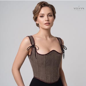 May include: A taupe-colored corset top featuring vertical paneling, a scoop neckline, and dark brown trim. The garment includes shoulder straps with black ribbon ties threaded through metal eyelets. The brand name VELVYN NOIR is visible.