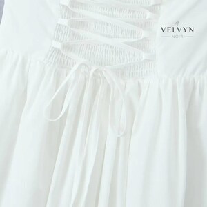 May include: Close-up of a white garment featuring a corset-style back with crisscross ribbon lacing. The fabric is gathered and flowing, with a soft texture. The brand name "VELVYN NOIR" is printed in black.