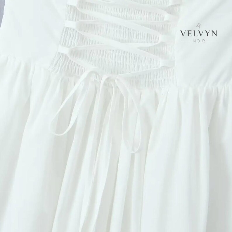 May include: Close-up of a white garment featuring a corset-style back with crisscross ribbon lacing. The fabric is gathered and flowing, with a soft texture. The brand name "VELVYN NOIR" is printed in black.