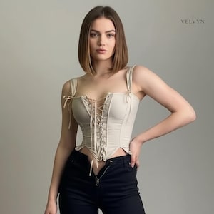 May include: A beige corset top with spaghetti straps and a lace-up front. The corset has a structured design and a slightly ruffled hem. The model is wearing black pants. The brand name "VELVYN" is visible in the top right corner.