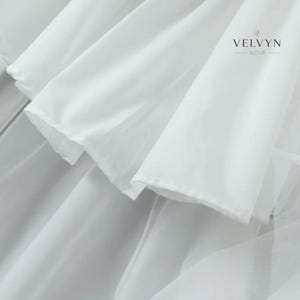 May include: Close-up of white fabric layers with a soft, flowing texture. The image features multiple layers of delicate, semi-transparent material, creating a sense of depth and movement. The brand name "VELVYN NOIR" is visible in the upper right corner.