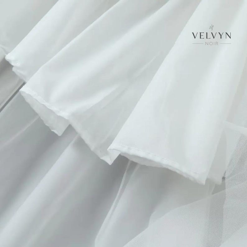 May include: Close-up of white fabric layers with a soft, flowing texture. The image features multiple layers of delicate, semi-transparent material, creating a sense of depth and movement. The brand name "VELVYN NOIR" is visible in the upper right corner.