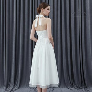 May include: A white halter dress with a bow at the neck and a lace-up back. The dress has a flowing skirt that falls to the mid-calf, with a sheer, layered hem. The dress is suitable for a wedding or formal event.