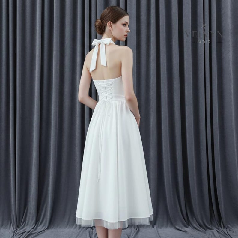 May include: A white halter dress with a bow at the neck and a lace-up back. The dress has a flowing skirt that falls to the mid-calf, with a sheer, layered hem. The dress is suitable for a wedding or formal event.