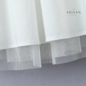 May include: Close-up of a white garment with a layered design. The top layer is a smooth, opaque fabric, while the bottom layer is a sheer, net-like material. The brand name "VELVYN NOIR" is visible in the upper right corner.