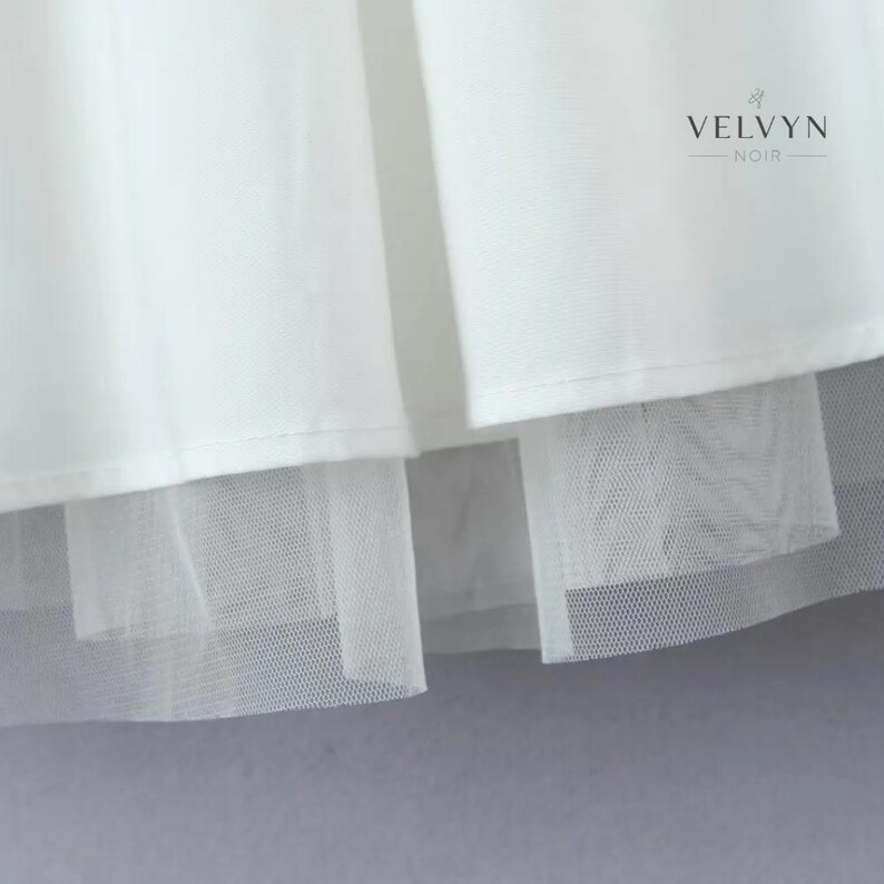 May include: Close-up of a white garment with a layered design. The top layer is a smooth, opaque fabric, while the bottom layer is a sheer, net-like material. The brand name "VELVYN NOIR" is visible in the upper right corner.
