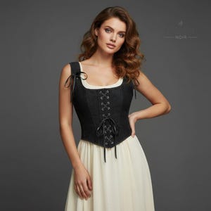 K&ouml;nnte beinhalten: Ein schwarzes Jeans-Korsett mit Schn&uuml;rung vorne und schwarzen B&auml;ndern. Das Korsett hat einen quadratischen Ausschnitt und wird mit einem cremefarbenen Kleid kombiniert. Das Bild zeigt den Text "NOIR" in der oberen rechten Ecke.