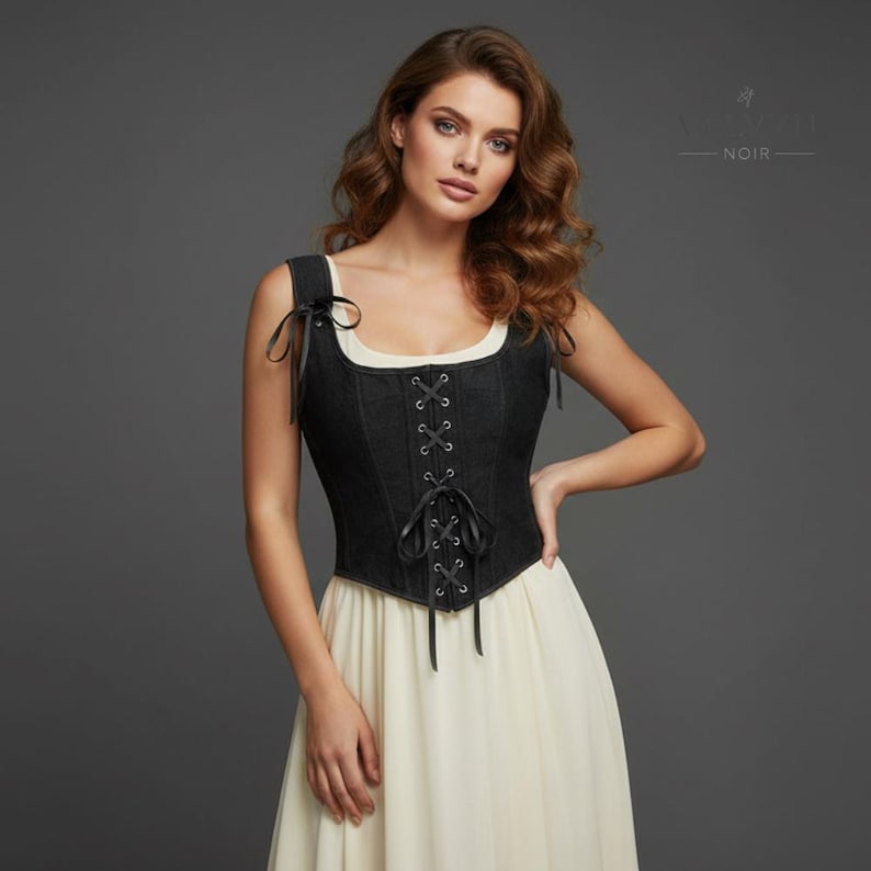 K&ouml;nnte beinhalten: Ein schwarzes Jeans-Korsett mit Schn&uuml;rung vorne und schwarzen B&auml;ndern. Das Korsett hat einen quadratischen Ausschnitt und wird mit einem cremefarbenen Kleid kombiniert. Das Bild zeigt den Text "NOIR" in der oberen rechten Ecke.