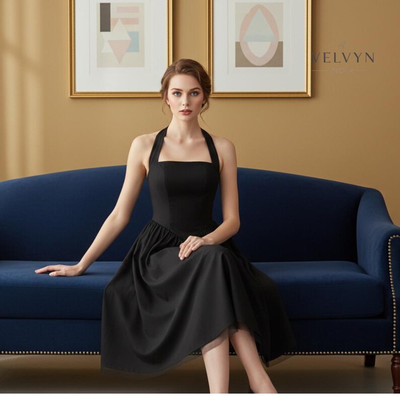 May include: A black halter dress with a square neckline and a flared skirt. The dress is knee-length and is worn by a person sitting on a blue sofa. The background features framed artwork and a gold-colored wall.