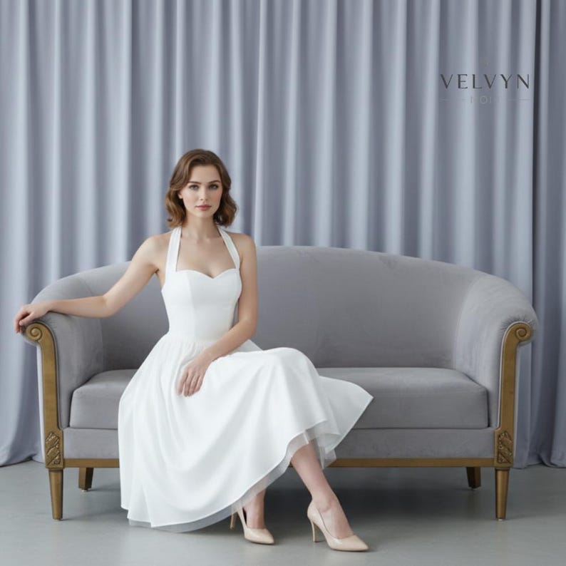 May include: A white halter dress with a sweetheart neckline and a flowing skirt. The dress is knee-length and paired with nude heels. The model is seated on a gray velvet sofa with gold trim. The background features gray curtains, and the brand name "VELVYN" is visible.