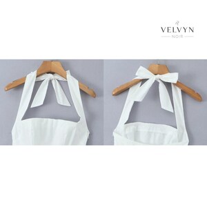 May include: White halter top with a bow detail at the back, displayed on a wooden hanger. The top has a square neckline and is made of a smooth fabric. The brand name "VELVYN NOIR" is visible in the top right corner.