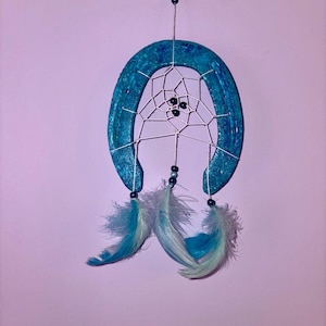 Peut inclure: Un attrape-rêves en forme de fer à cheval turquoise avec un motif de toile d'araignée blanche, des perles noires et des plumes turquoise et blanches. L'attrape-rêves est suspendu sur un fond violet clair.
