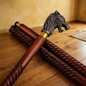 Puede incluir: Un bastón con un eje de madera marrón oscuro con una textura de espiral retorcida y un mango de metal con forma de cabeza de lobo. El mango tiene un acabado en tono bronce oscuro y está unido al eje mediante un collar de metal dorado pulido.