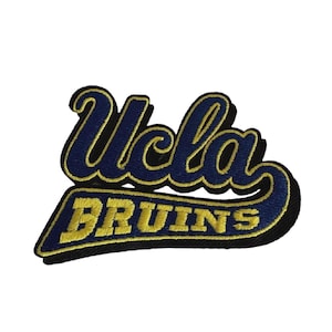 Puede incluir: Parche bordado con el texto "UCLA BRUINS" en azul marino y dorado. La palabra "UCLA" está en cursiva, y "BRUINS" está en negrita. El parche tiene un borde negro y está sobre un fondo blanco.