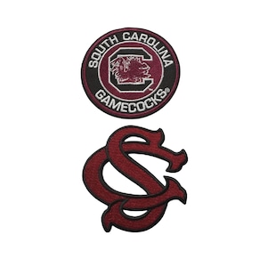 Puede incluir: Dos parches bordados. El parche superior es circular con el texto "SOUTH CAROLINA GAMECOCKS" alrededor del borde y un gallo en el centro. El parche inferior es un "SC" estilizado en granate.