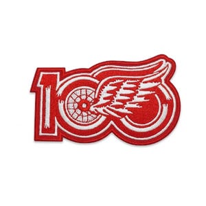 May include: Red and white embroidered patch featuring the number "100" and a stylized winged wheel logo. The patch is a commemorative item, likely for a sports team or organization.