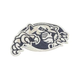 Pode incluir: Patch bordado com um puma estilizado em azul marinho e branco sujo. O puma é retratado numa pose dinâmica, com as garras estendidas. O patch tem uma borda branca e é adequado para roupas ou acessórios.