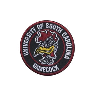 Puede incluir: Un parche redondo con el logotipo de Gamecock de la Universidad de Carolina del Sur. El parche tiene un fondo negro con un borde rojo y texto blanco que dice "UNIVERSITY OF SOUTH CAROLINA" y "GAMECOCK". La mascota Gamecock está en el centro.