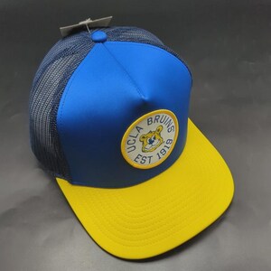 以下が含まれることがあります： メッシュバックの青と黄色のトラッカーハット。フロントパネルは青色で、つばは黄色です。「UCLA Bruins Est 1919」の文字と漫画のクマが描かれた円形のパッチが付いています。