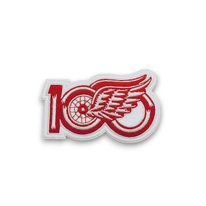 May include: Embroidered patch featuring a red and white design. The patch displays the number "100" with a stylized wheel and wing motif. The design is outlined in white, creating a contrasting border. This patch is suitable for apparel and accessories.
