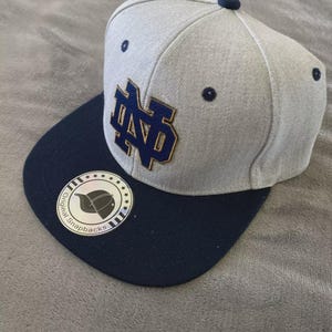 May include: A gray and navy blue snapback hat with the letters "ND" in a gold and navy blue design. The hat has a flat brim and a round sticker on the brim. The hat is on a gray surface.