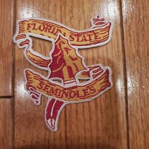 Può includere: Toppa ricamata con le parole "FLORIDA STATE SEMINOLES" in lettere dorate su un disegno rosso e oro. La toppa ha un design stilizzato a lancia e banner, con un bordo bianco. La toppa è posizionata su una superficie di legno.