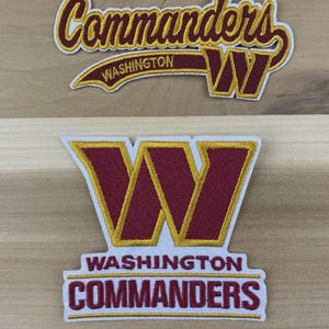May include: Two embroidered patches featuring the Washington Commanders logo. One patch has the word "Commanders" in a script font above a "W" logo, with "Washington" below. The other patch displays the "W" logo above "Washington Commanders".