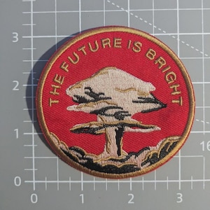 May include: Circular embroidered patch with the gold text "THE FUTURE IS BRIGHT" on a red backdrop. It features a beige and black stylized tree design, with gold detailing. The patch is approximately 7.6 cm in diameter.