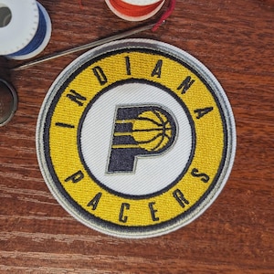 Könnte beinhalten: Gestickter Aufnäher mit dem Logo der Indiana Pacers. Der Aufnäher ist kreisförmig, mit einem gelben Außenring und dem Teamnamen in Schwarz. Die Mitte hat einen weißen Hintergrund mit dem Logo des Teams, einem stilisierten 'P' mit Basketball.