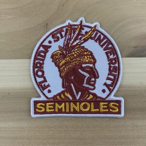 Può includere: Toppa ricamata con il logo dei Seminoles della Florida State University. La toppa ha uno sfondo bianco con un disegno di testa di nativo americano marrone e oro. Le parole "FLORIDA STATE UNIVERSITY" e "SEMINOLES" sono ricamate.
