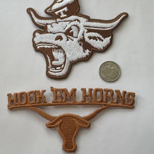 Könnte beinhalten: Gestickte Aufnäher mit einem Longhorn-Logo und dem Schriftzug "Hook Em Horns". Die Aufnäher sind in Braun- und Weißtönen gehalten. Eine silberne Münze ist ebenfalls vorhanden.