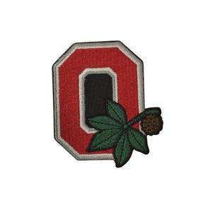 Può includere: Toppa ricamata con la lettera "O" rossa, interno nero e bordo bianco. Una foglia verde e una noce marrone sono attaccate. La toppa è probabilmente per abbigliamento o accessori.