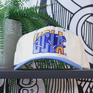 以下が含まれることがあります： 青い縁と青とオレンジのグラフィックが付いた白い野球帽。グラフィックは、クマのイラストとともに「UCLA」の文字を特徴としています。帽子は黒い棚に飾られています。