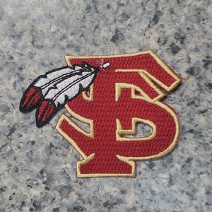 Può includere: Toppa ricamata con un design intrecciato rosso e oro "F" e "S", e un'emblema di piuma. La toppa ha uno sfondo rosso con finiture dorate e un contorno nero. La piuma presenta dettagli rossi, bianchi e neri.