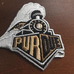 May include: Embroidered patch with the word "PURDUE" in gold and black, and a train design. The patch has a white border and a "TM" symbol, suitable for clothing or accessories.