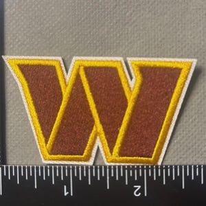 May include: Embroidered patch featuring a maroon "W" with a gold outline and white border. The patch is approximately 2 inches wide and 2 inches tall. The patch is on a gray background.