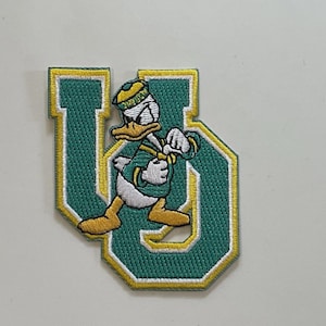 May include: Embroidered patch featuring Donald Duck in a green and gold uniform, running across a green "U" shape. The patch has a white border and is likely for apparel or accessories.
