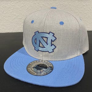 May include: A grey and light blue snapback cap featuring the interlocking "NC" logo in light blue. The cap has a flat peak and a structured crown. The cap is a sports-themed accessory.