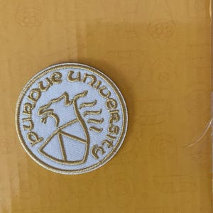 May include: A circular embroidered patch with the words "Purdue University" in gold thread. The patch has a white background, a gold outline, and a gold graphic of a stylized Boilermaker Special.
