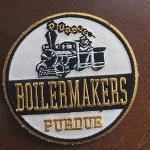 May include: Embroidered patch with a black and white train design on a white background. The patch features the words "BOILERMAKERS" and "PURDUE" in gold lettering, with a gold border. A train driver is visible.