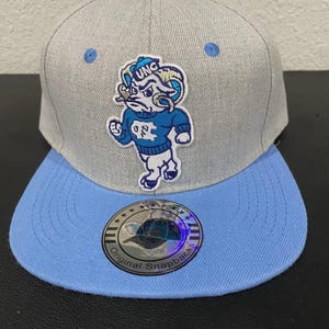 May include: A grey and light blue snapback cap with a cartoon ram mascot wearing a blue sweater with "UNC" on it. The cap has a light blue brim and an "Original Snapbacks" sticker.