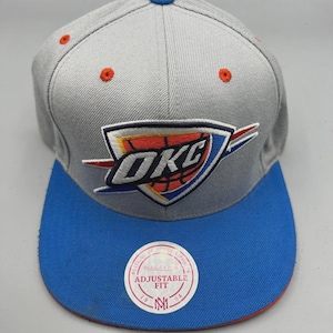May include: A gray and blue adjustable fit baseball cap with the OKC logo. The cap features a flat blue brim, gray crown, and orange accents. The OKC logo is embroidered on the front.
