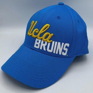以下が含まれることがあります： 青い野球帽。黄色と白で「UCLA BRUINS」の文字が書かれています。帽子はカーブしたつばと構造化されたクラウンが付いています。帽子は丈夫な生地で作られています。
