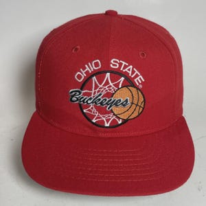 May include: Red baseball cap with a flat brim. The cap features the words "OHIO STATE" and "Buckeyes" embroidered in white and black, along with a basketball graphic in orange and white.